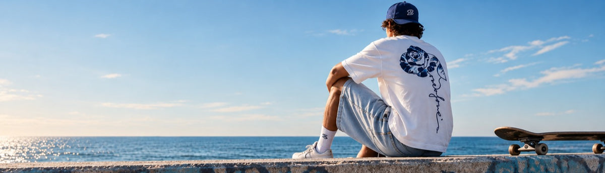 Person sitzt mit dem Ruecken zur Kamera auf einer Ufermauer am Meer, traegt das weisse oneofone-Shirt mit Rosenstickerei und die Rose Denim Jorts, Skateboard liegt daneben.